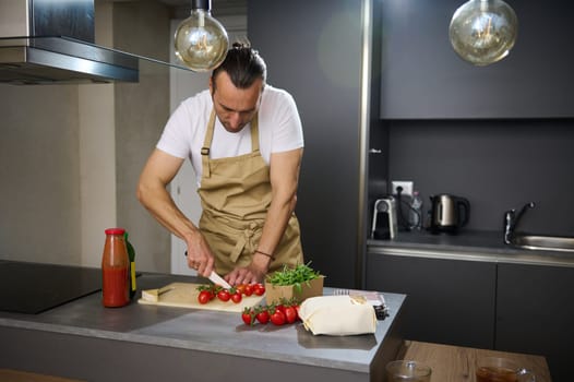 Handsome young man in beige chef's apron, cooking healthy vegetarian salad in the modern minimalist home kitchen, standing at kitchen island and chopping fresh ripe cherry tomatoes. People. Culinary