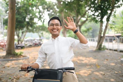 Cheerful asian businessman walking with his bicycle and waving hand for greetings, saying hello.
