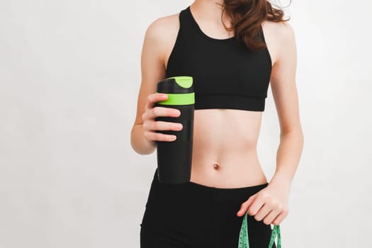 Young beautiful caucasian teenage girl in a fitness suit holds a measuring tape and a bottle with a sports drink in her hands on the right against a white wall with copy space on the left, close-up side view. The concept of sport, fitness and modern lifestyle.