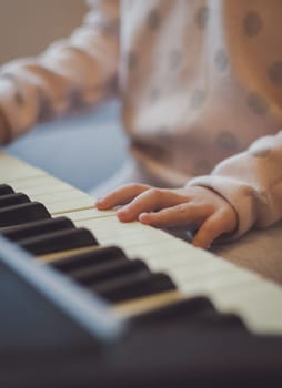 The hands of a little caucasian girl presses the keys on an electric piano while sitting on a sofa in the room, close-up side view with depth of field. Music education concept.