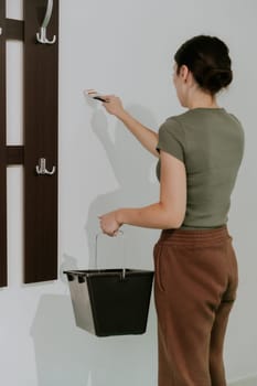 One young beautiful Caucasian brunette stands sideways, holds a bucket of paint and paints the wall with white paint with a brush near the wardrobe, close-up side view.