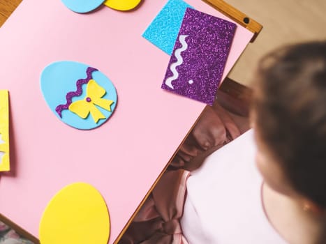 Felt blue egg with a yellow bow, lilac zigzag and a set of stickers on a pink background and a little caucasian girl sitting at a children's table in her room, close-up flat lay. Concept diy, crafts, needlework, children art, artisanal, creative children.