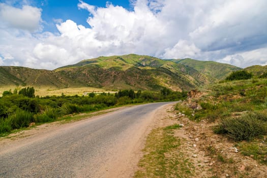 Asphalt road in the countryside with mountains towering in the background, under a clear blue sky with fluffy clouds