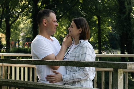 Portrait of a young beautiful Caucasian couple standing and looking at each other in love on an old wooden bridge over a brook in the park on a summer day...