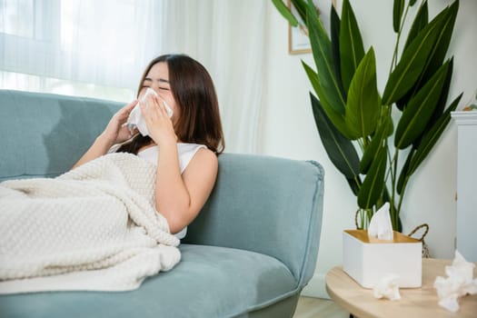 Ill Asian young woman cold covered with blanket sitting on sofa and sneeze with tissue paper at home, Female sick allergic blowing nose sneezing in tissue at home, Flu health care