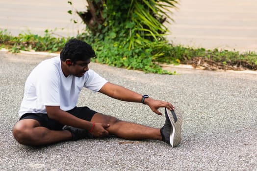 Close up Asian young athlete sport runner black man wear watch he sitting pull toe feet stretching legs and knee before running at outdoor street health park, healthy exercise before workout concept