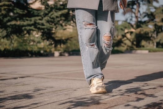 A woman is walking down a sidewalk wearing ripped jeans and a gray jacket. The image conveys a casual and relaxed atmosphere, as the woman is enjoying her walk