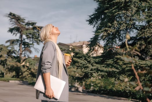 A woman is walking down the street with a white laptop bag and a cup of coffee. She is smiling and enjoying the sunny day