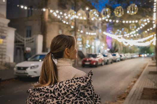 A woman wearing a leopard print coat stands on a street at night. The street is lit up with Christmas lights, creating a festive atmosphere. There are several cars parked along the street