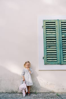 Little girl stands holding a pink toy rabbit by the paw, leaning on the wall of an old house. High quality photo