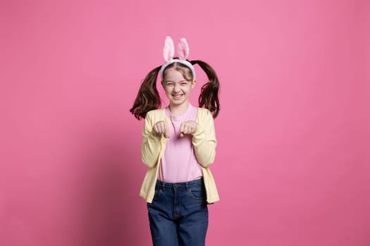 Adorable little girl spending Easter holiday by hopping around like a rabbit for the camera while wearing bunny ears. Delighted happy youngster playing and chuckling on pink background.