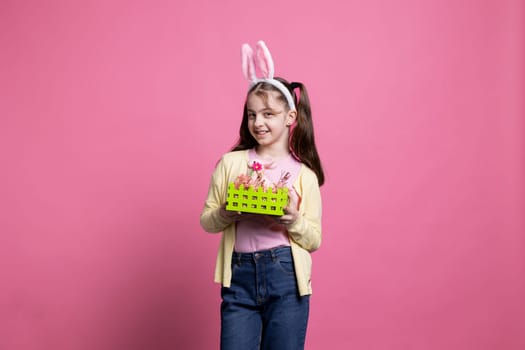 Happy confident girl with bunny ears presenting a basket filled with painted handmade easter decorations over pink background. Young cheerful kid showing festive colorful ornaments in studio.