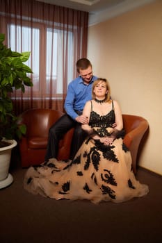 A plump woman in royal dress and a skinny man together in honeymoon hotel room. An adult newlywed couple embraces in an intimate setting. Queen Girl and guy in love