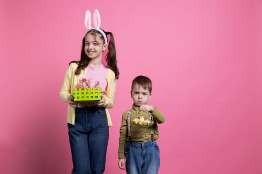 Young kids holding handmade decorations and ornaments on camera, posing for an easter celebration photoshoot. Adorable boy and girl feeling joyful about spring celebration event in studio.