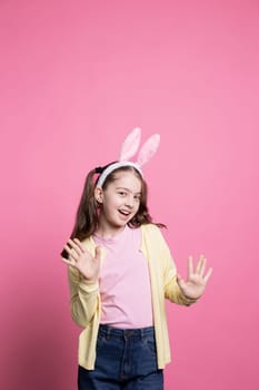 Little child acting excited and cheerful about easter festivity, posing with confidence against pink background. Young small kid wearing bunny ears and pigtails, feeling cheerful and carefree.