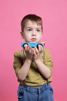 Toddler playing with a blue automobile toy in front of the camera, satisfied child having fun against pink background. Adorable kid loving play with brightly colored vehicle in studio.