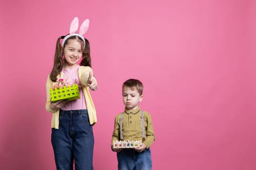 Joyful siblings feeling happy celebrating easter holiday and spring time, little girl giving thumbs up on camera. Brother and sister showing baskets filled with painted eggs and decorations.