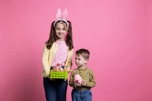 Playful children showing painted eggs and decorating for easter celebration festivity, standing together over pink background. Brother and sister feeling cheerful and enthusiastic about april event.