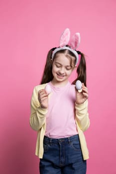 Small child posing with confidence in front of camera, showing her painted colorful eggs for easter celebration. Young sweet kid with bunny ears smiling in studio, presenting handmade decorations.