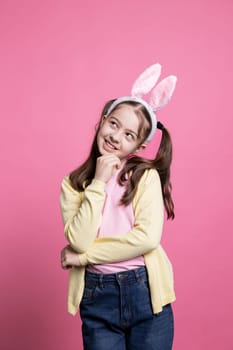 Little toddler smiling in studio thinking about presents, being excited about easter holiday celebration. Joyful schoolgirl with bunny ears and pigtails being pensive on camera, sweet kid.