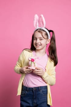 Cheerful small kid posing with a pink rabbit toy on camera, wearing fluffy bunny ears and holding easter ornaments over background. Young toddler with pigtails being excited about festive event.