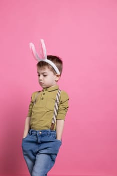 Lovely small boy wearing fluffy bunny ears and posing with confidence on camera, feeling excited about easter celebration and standing against pink backdrop. Cheerful preschooler with adorable outfit.