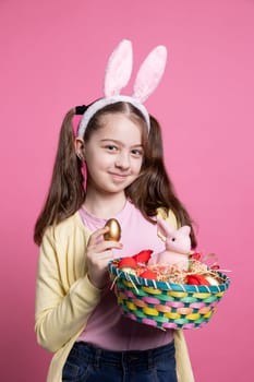 Smiling cute little girl showing colorful eggs and rabbit toy in an arrangement, posing with confidence in pink studio. Young child with bunny ears holding a basket with lovely decorations.