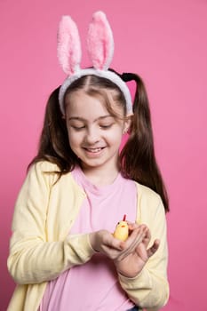 Small young kid showing a cute stuffed chick toy in studio, joyful little girl with golden arrangement over pink background. Youngster with sweet fluffy ears for easter festivity in front of camera.