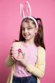 Happy positive girl with bunny ears holds a pink easter egg in studio, feeling proud about her festive decorations handmade. Small child being confident on camera, spring holiday event.