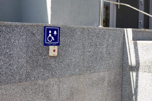 Access to building door for disabled people. Sign with braille text and symbol of person in wheelchair near button to call for help for climbing stairs