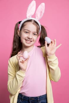 Young cute girl posing with confidence in front of camera, presenting her painted pink eggs for easter festivity. Little schoolgirl wearing bunny ears smiling in studio, shows handmade decorations.