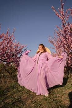 Woman blooming peach orchard. Against the backdrop of a picturesque peach orchard, a woman in a long pink dress and hat enjoys a peaceful walk in the park, surrounded by the beauty of nature