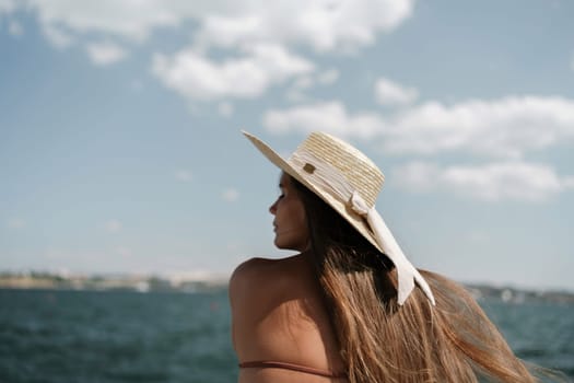 A woman in a swimsuit sits with her back holding a hat, looks at the ocean, sunny day, relaxes