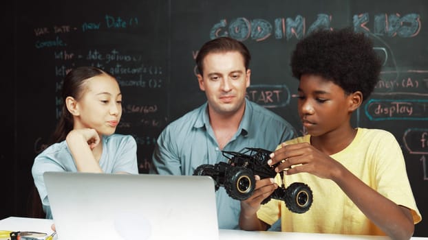 Smart teacher inspect car model construction to diverse student. Highschool children with mixed races listening mentor explain robotic model system at table with laptop and wires placed. Edification.