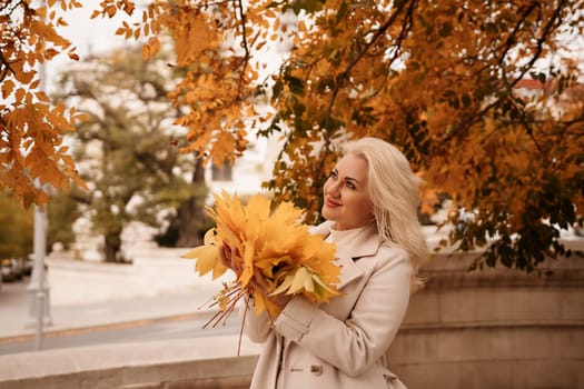 A blonde woman is holding a bunch of yellow leaves in her hand. She is standing in front of a tree with leaves that are changing colors. The scene has a peaceful and serene mood