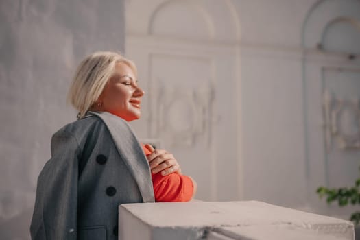 A woman is standing in front of a white building, wearing a gray jacket and a red shirt. She is smiling and looking at the camera