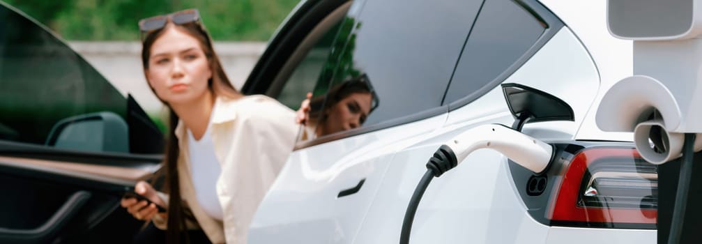 Young woman use smartphone to pay for electricity at public EV car charging station green city park. Modern environmental and sustainable urban lifestyle with EV vehicle. Panorama Expedient