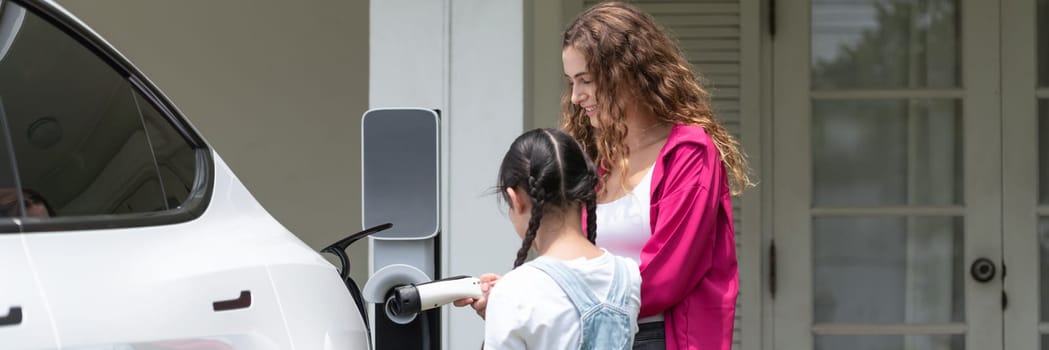 Happy little young girl learn about eco-friendly and energy sustainability as she help her mother recharge electric vehicle from home EV charging station. EV car and modern family. Panorama Synchronos