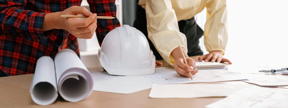 Professional engineer draws a blueprint on meeting table with blueprint and equipment scattered around while project manager using laptop analysis data at meeting room. Closeup. Delineation.