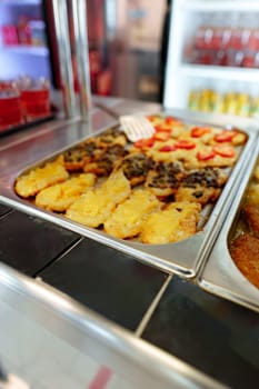 A bustling cafeteria serving line is filled with an array of dishes during a busy lunchtime. Trays of hot food are neatly arranged under the protective glass shield, with customers in the background selecting their meal choices. The variety suggests a diverse menu, catering to different tastes.