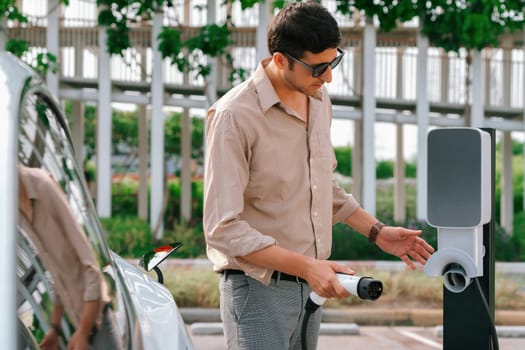 Young man put EV charger to recharge electric car's battery from charging station in city commercial parking lot. Rechargeable EV car for sustainable environmental friendly urban travel. Expedient