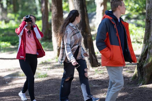 A group of friends, casually dressed, walking through a dense forest with tall trees and sunlight peeking through the canopy. The individuals are engaged in conversation as they trek along a dirt path.