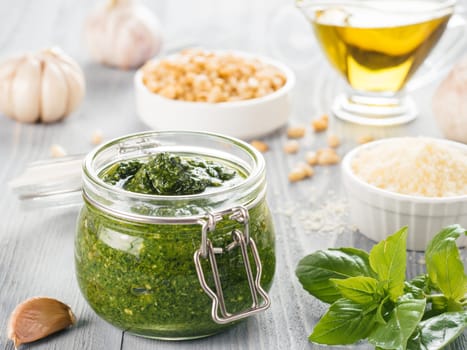 Homemade pesto sauce and ingredients on gray wooden background. Close up wiev of basil pesto in glass jar with ingredients.