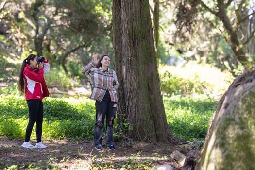 Two female friends standing side by side next to a tall tree in an outdoor setting. Both women are casually dressed and appear to be engaged in conversation or enjoying the surroundings.