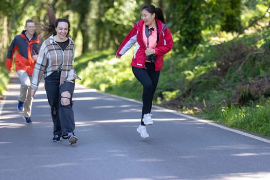 Three young girls, friends, are running enthusiastically down a dusty road surrounded by dense trees in a forest setting. The girls are wearing casual clothing and appear to be enjoying their outdoor activity.