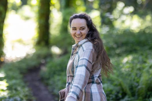 A woman is walking down a narrow path in a dense forest, surrounded by tall trees and lush greenery. The path appears well-worn and leads deeper into the woods.