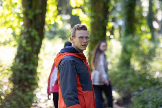 A man wearing a red and black jacket is walking through a dense forest. The trees around him are tall and leafy, casting shadows on the forest floor as he moves forward.