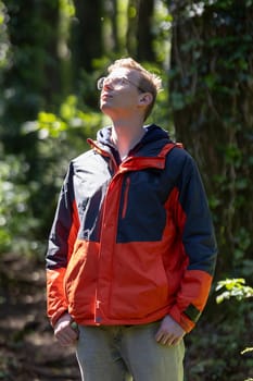 A man is standing alone in a forest, gazing upwards towards the sky, surrounded by tall trees and the sounds of nature.