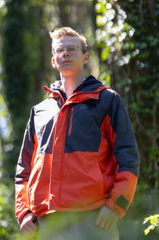 A man wearing a red and blue jacket is standing among the trees in a forest. He appears to be alone, surrounded by the natural environment.