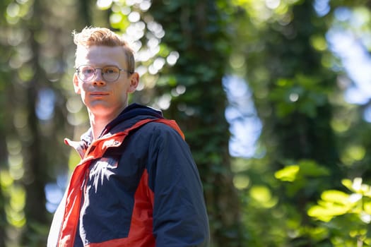 A man wearing glasses stands alone among the trees in a forest, surrounded by dense foliage and tall trunks. He gazes around, taking in the natural surroundings.